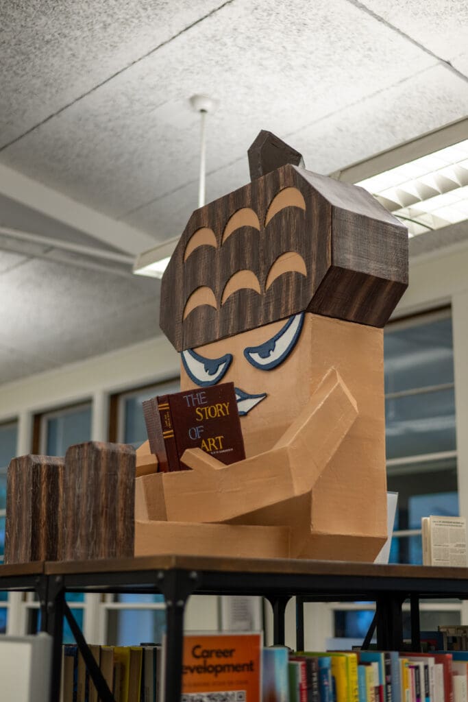 A large, stylized wooden sculpture of Menlo College’s Oakie mascot sits on top of a bookshelf in Bowman Library, posed reading a book titled “The Story of Art.” The sculpture has carved wood-grain textures, expressive eyes, and oversized blocky hands and legs. Library shelves and fluorescent lights are visible in the background.