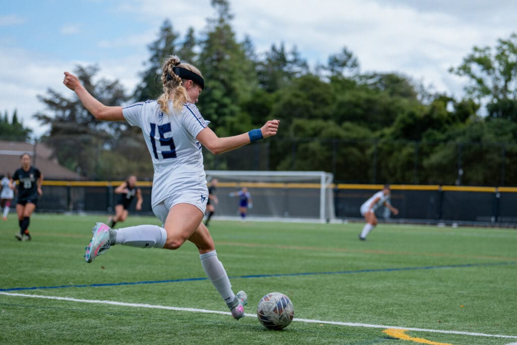 A female soccer player in a white uniform, number 15, attempts a kick on a grassy field. Teammates and opponents are in the background. Overcast skies.
