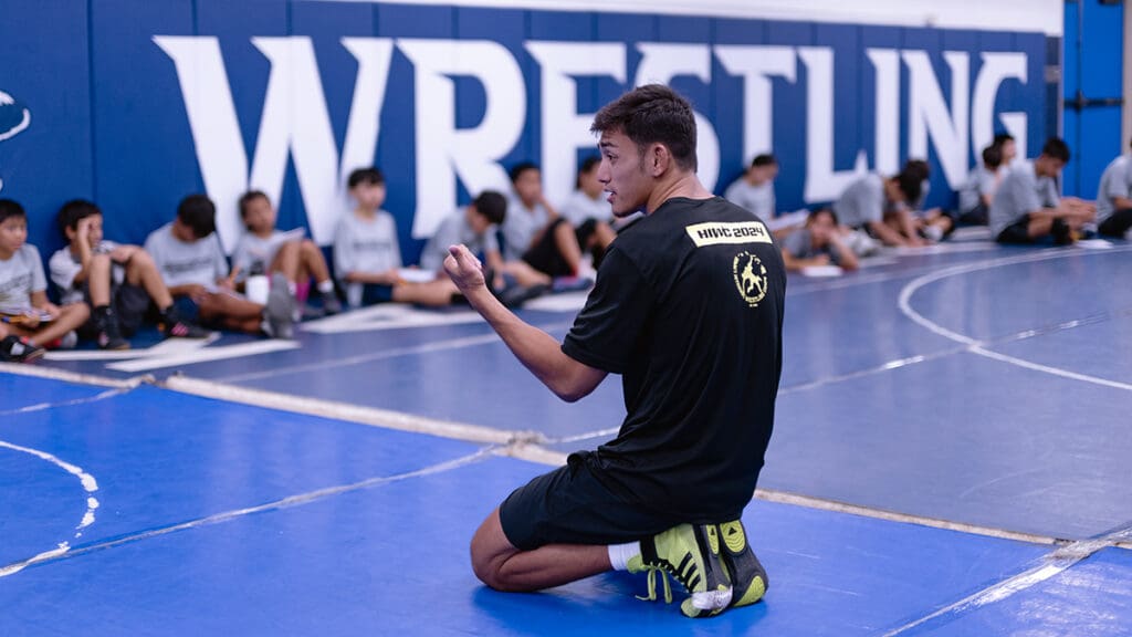 A wrestling coach kneels on a blue mat, gesturing as he instructs a group of seated young athletes in a gym, with "Wrestling" on the wall behind.