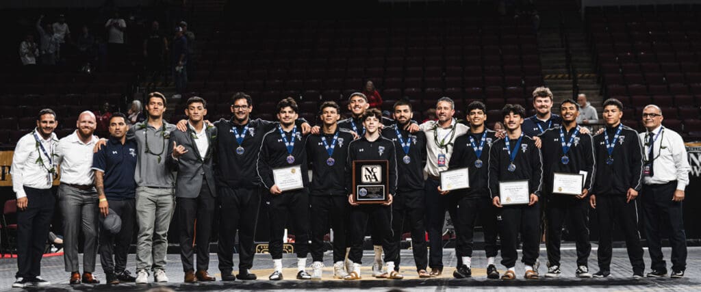 A group of 15 men stand in a row, all smiling. They wear matching black tracksuits with medals around their necks, holding certificates and a plaque, on a stage in an arena.
