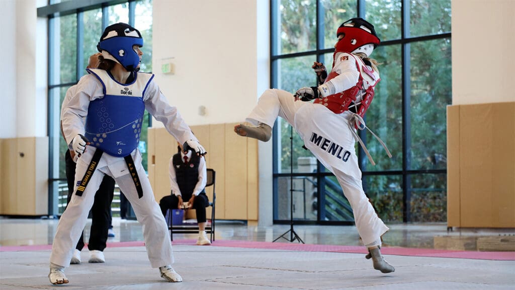 Two taekwondo athletes in protective gear are competing indoors. One delivers a high kick, while the other prepares to block. A referee watches attentively.
