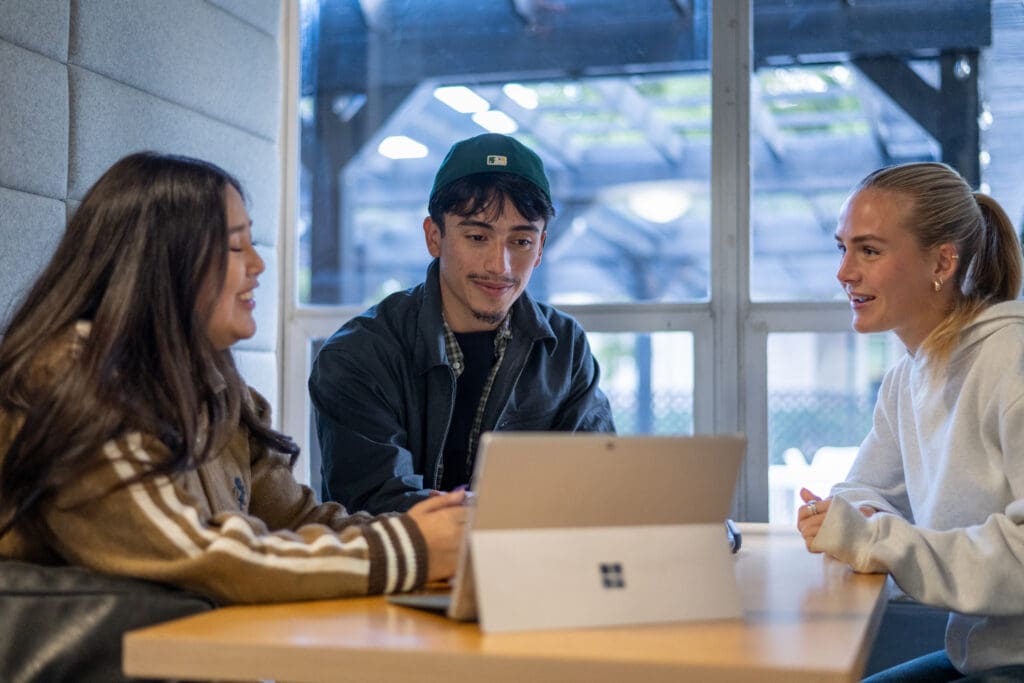 Three people sit around a table, engaged in conversation, with a laptop open. They appear relaxed and focused in a bright, modern setting.