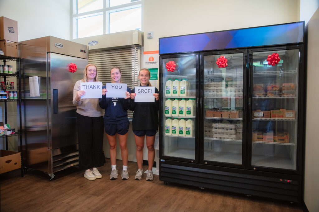 Three people stand indoors holding signs that read "THANK YOU SRCF!" in front of commercial refrigerators stocked with milk and other food items.