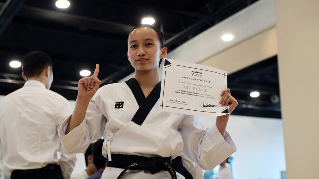 A person in a martial arts uniform holds a 1st place certificate, raising one finger in triumph. The setting is indoors with bystanders nearby.
