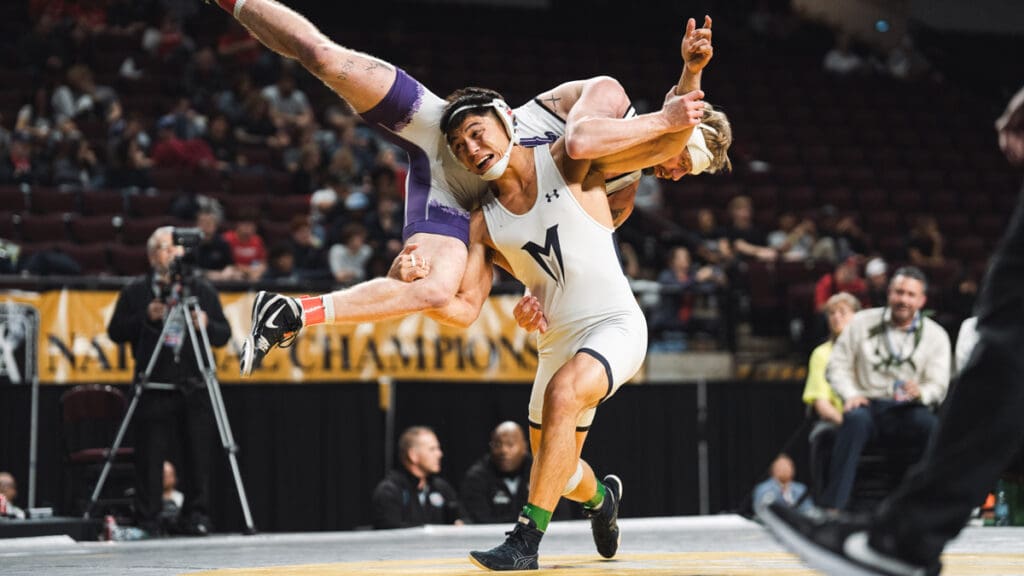 Two wrestlers in intense action on a mat; one athlete lifts the other off the ground. Audience and competition banner visible in the background.