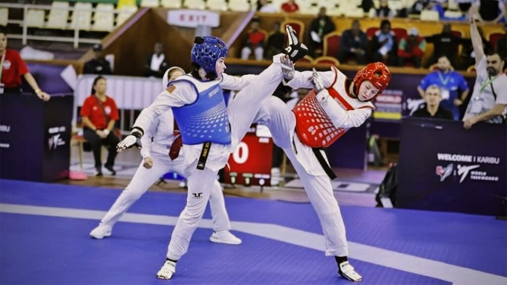 Two taekwondo athletes in protective gear compete intensely on a mat. One executes a high kick toward the other's head. A focused crowd watches closely.