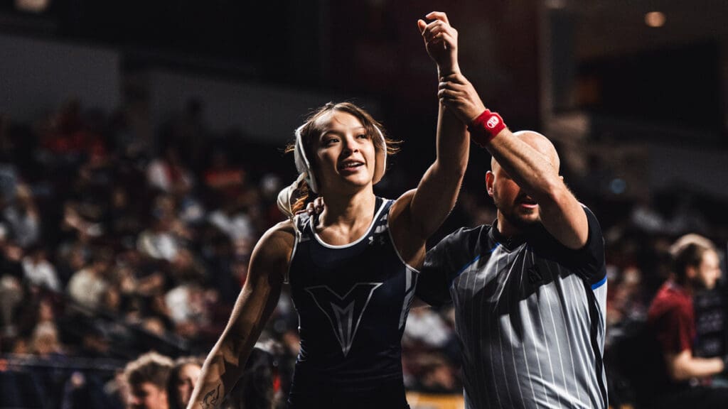 A victorious female wrestler in a dark uniform smiles as a referee raises her arm in triumph at a sports event, with a blurred cheering crowd.
