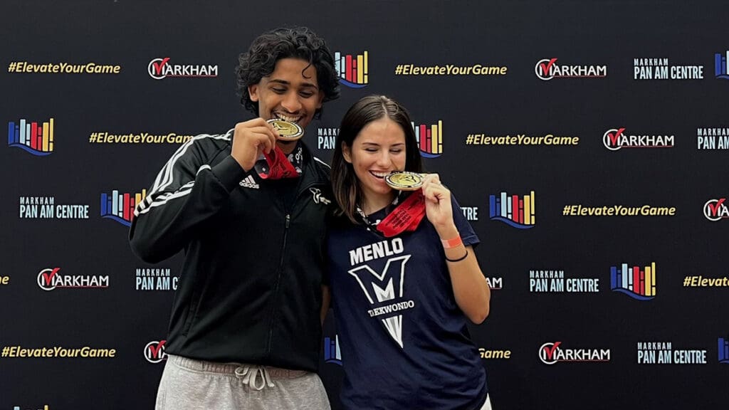 Two people smiling and biting gold medals, standing in front of a backdrop with "Markham Pan Am Centre" and "#ElevateYourGame" logos.