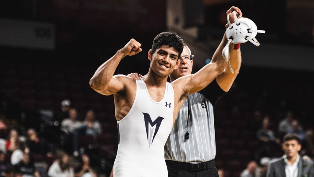 A triumphant wrestler in a white singlet with a blue "M" raises his fists in victory as a referee holds his arm. The background shows an audience in a sports arena.