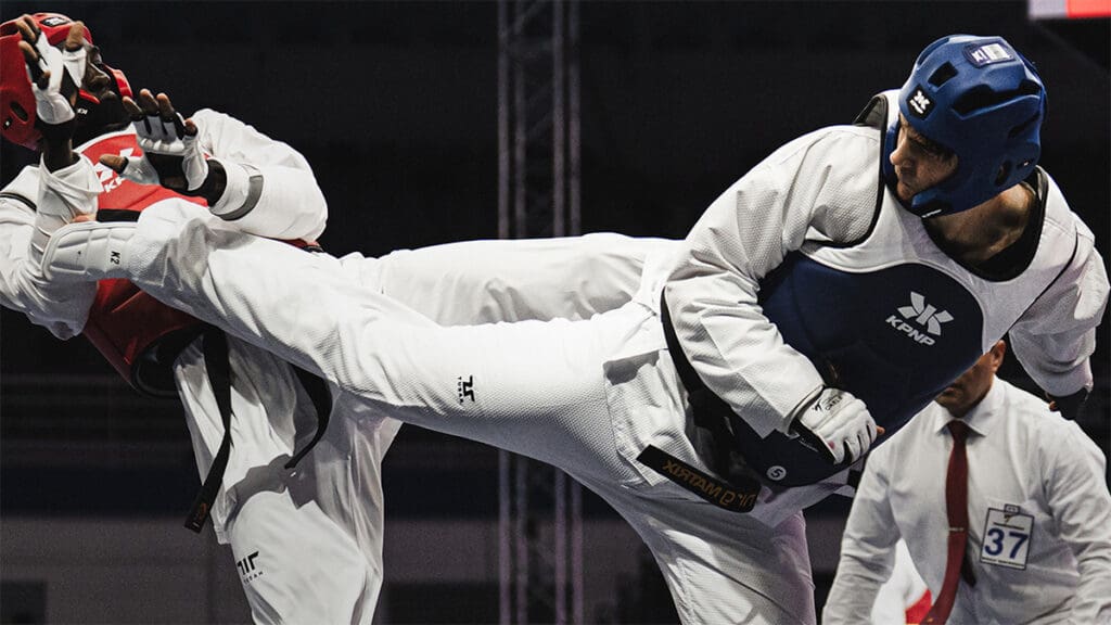 Two athletes in protective gear compete in a taekwondo match. One executes a high kick, the other guards. An official is seen in the background.