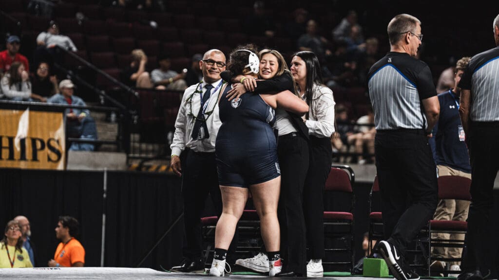 A female wrestler in a navy singlet hugs a coach on a platform, surrounded by officials. The crowd in the dimly lit arena watches, creating a celebratory tone.