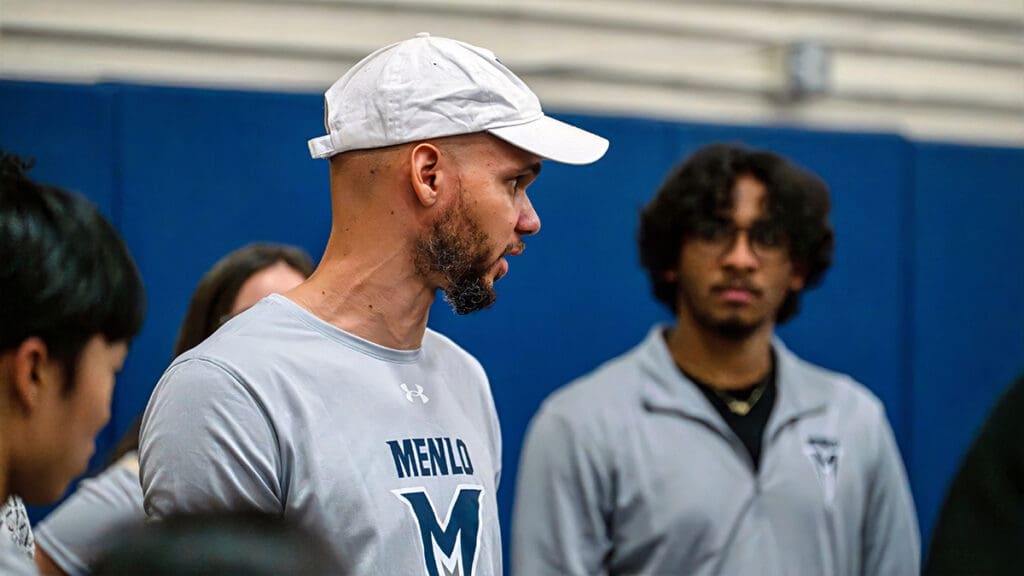 A man in a white cap and "Menlo" shirt speaks to a group in a gym. Another person listens intently, conveying a focused and attentive atmosphere.