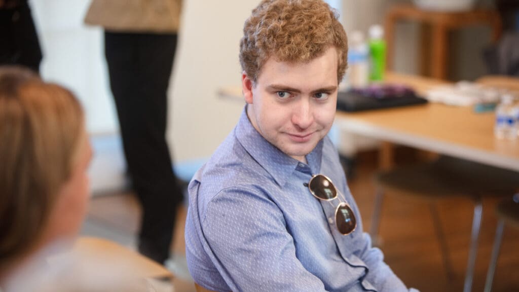 A person with curly hair and a blue shirt sits in a casual setting, holding sunglasses and smiling. An engaging conversation appears to be in progress.