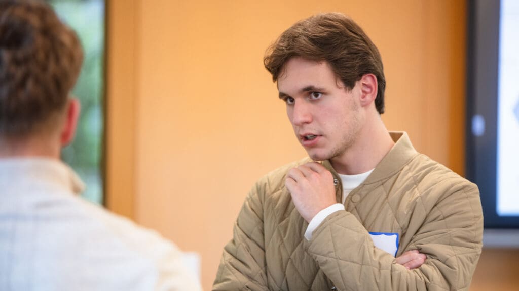 Young man in a quilted jacket thoughtfully listens in a conversation with another person in a bright room, evoking a focused and engaged mood.