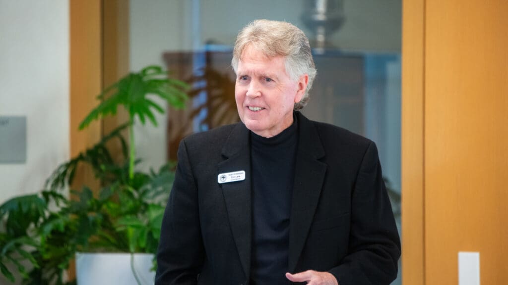 An older man in a black blazer smiles warmly in an office setting. He wears a name tag and stands near green potted plants and wooden paneling.