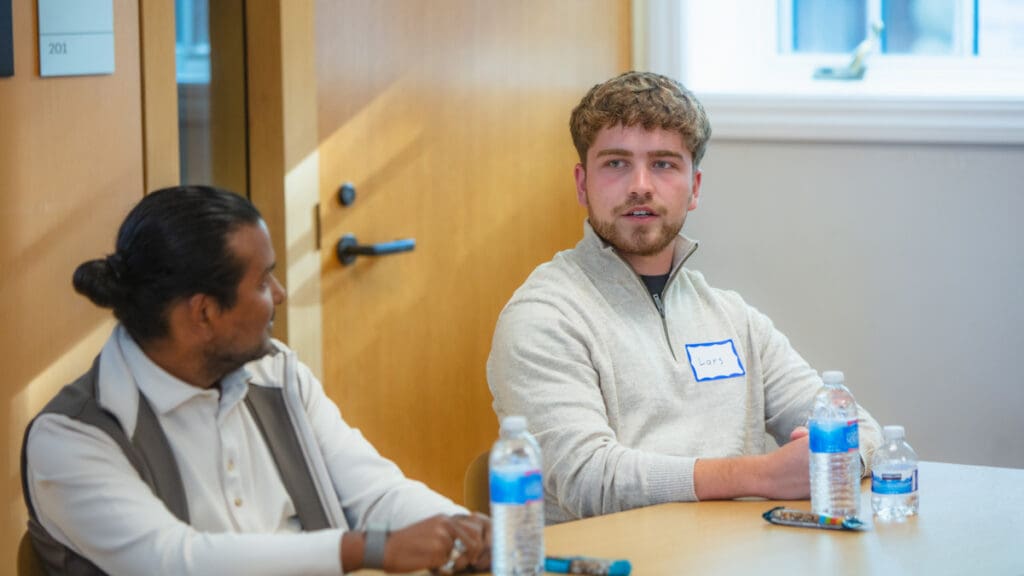 Two men are sitting at a table in a meeting room, engaged in conversation. They have water bottles in front of them. The atmosphere appears professional and focused.