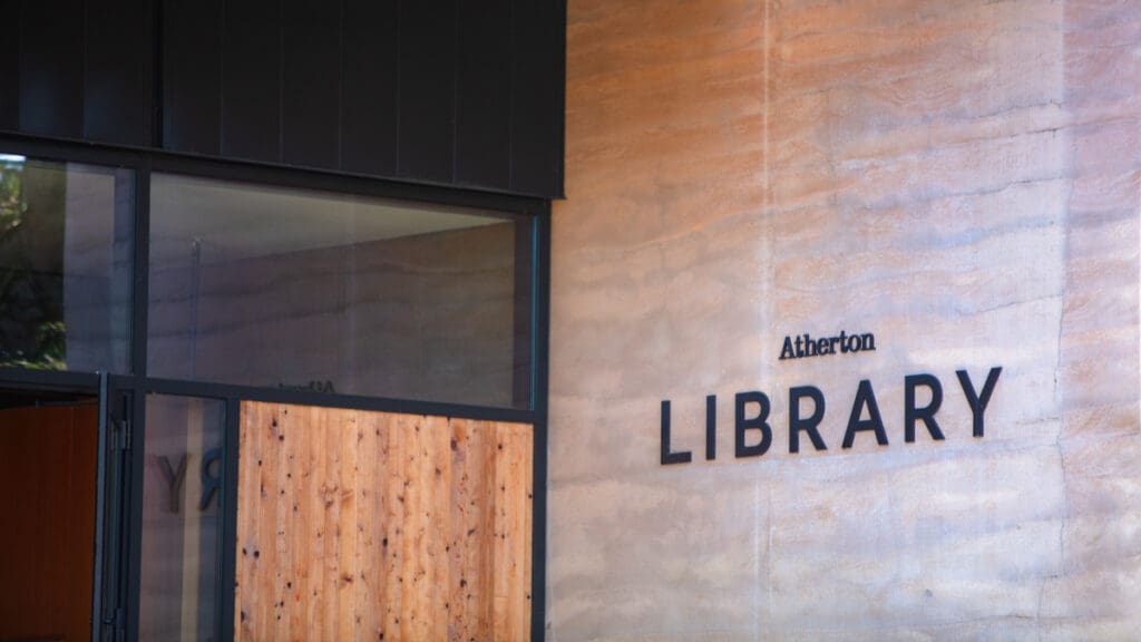 Modern library entrance with a wood and glass door, concrete walls, and "Atherton Library" text. Sleek design, conveying a welcoming atmosphere.