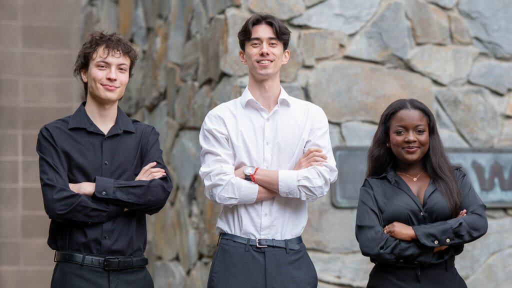 Three people stand confidently with arms crossed, smiling. They wear business attire. A stone wall serves as the backdrop, creating a professional tone.