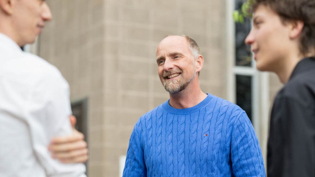 A smiling man in a blue sweater engages in conversation with two others outside. The mood is friendly and relaxed, with a blurred building background.