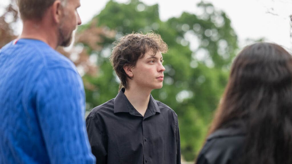 A young person in a black shirt stands outdoors, looking contemplative. Two others, partially visible, face them. Green trees form a blurred background.
