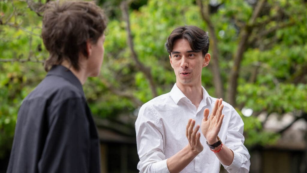 Two people engaged in conversation outdoors, with one clapping hands for emphasis. They stand against a backdrop of lush green trees, conveying a lively discussion.