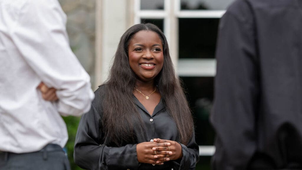 A woman in a black blouse smiles warmly while conversing with two people outdoors. The mood is friendly and engaging, suggesting a pleasant interaction.