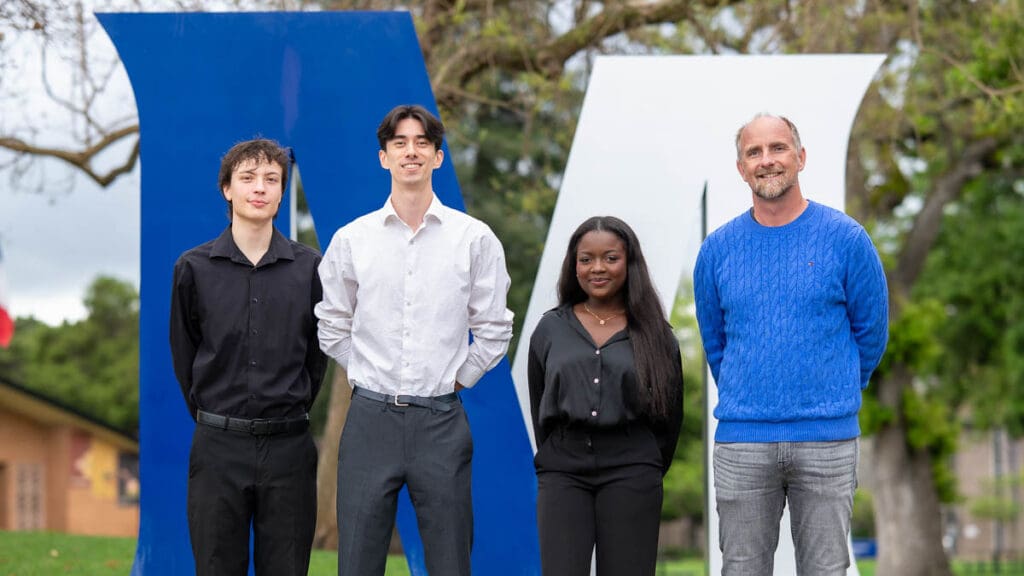 Four people stand in front of large blue and white letters outdoors. They smile warmly, conveying a friendly and welcoming atmosphere.