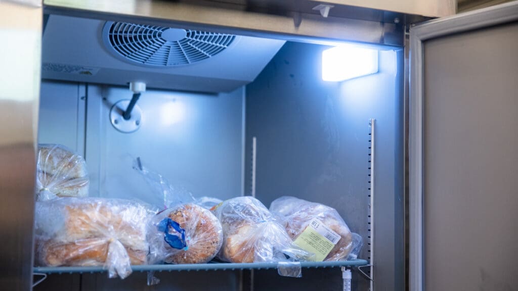 An open refrigerator with packaged bread on a metal shelf, illuminated by a bright interior light. The scene conveys a sense of freshness and organization.