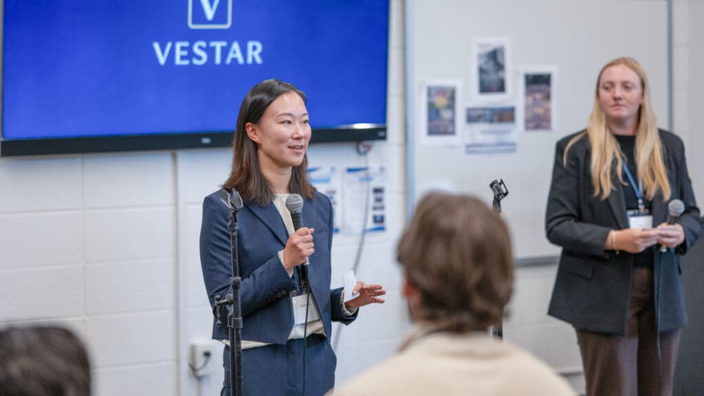A woman in a blue suit speaks into a microphone at a Vestar presentation. Another woman with blonde hair stands behind her, also holding a mic.