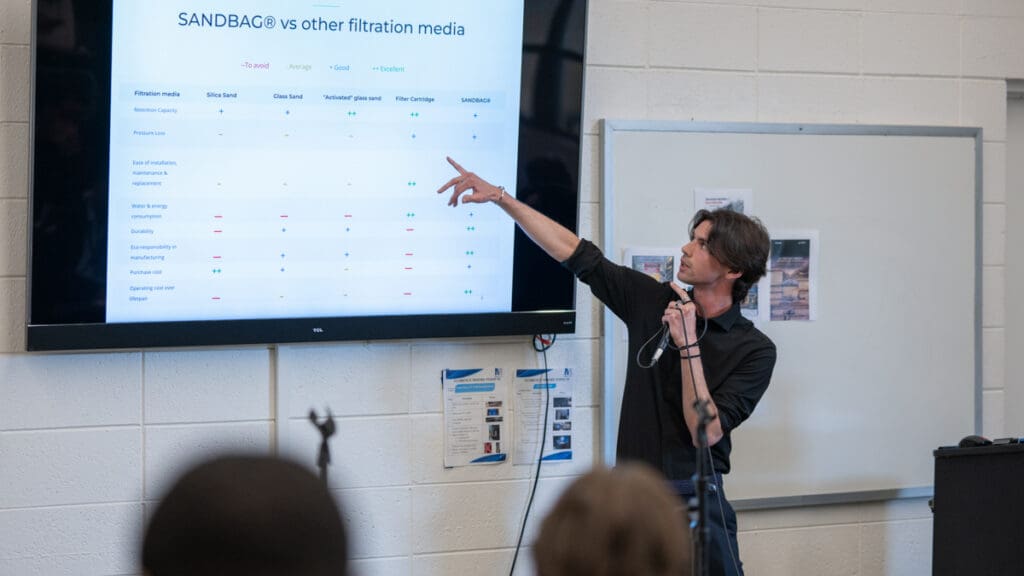 Man presenting in a classroom, pointing at a screen comparing filtration media. He holds a microphone, conveying focus and engagement.