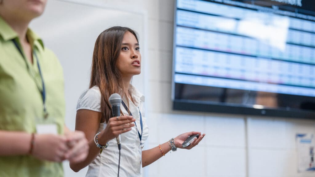 A woman in a white shirt speaks into a microphone, presenting in front of a large screen displaying data. She appears focused and confident.
