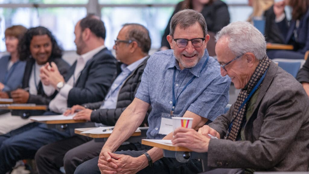 A group of diverse people seated in an auditorium, smiling and engaged in lively conversation. The atmosphere is warm and collegial.