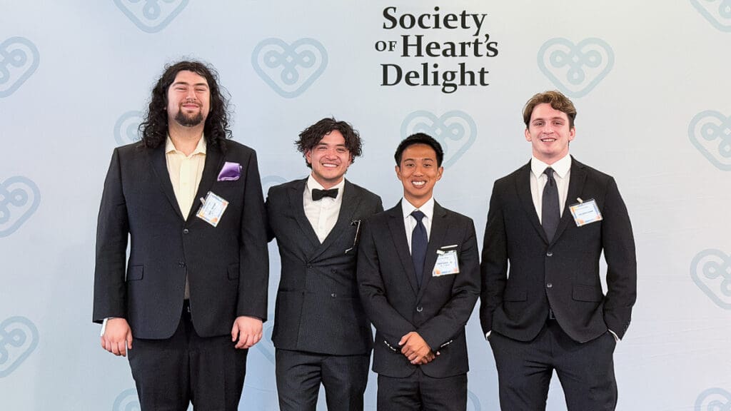 Three men in suits stand smiling in front of a backdrop with "Society of Heart’s Delight" logo. The mood is formal and cheerful.