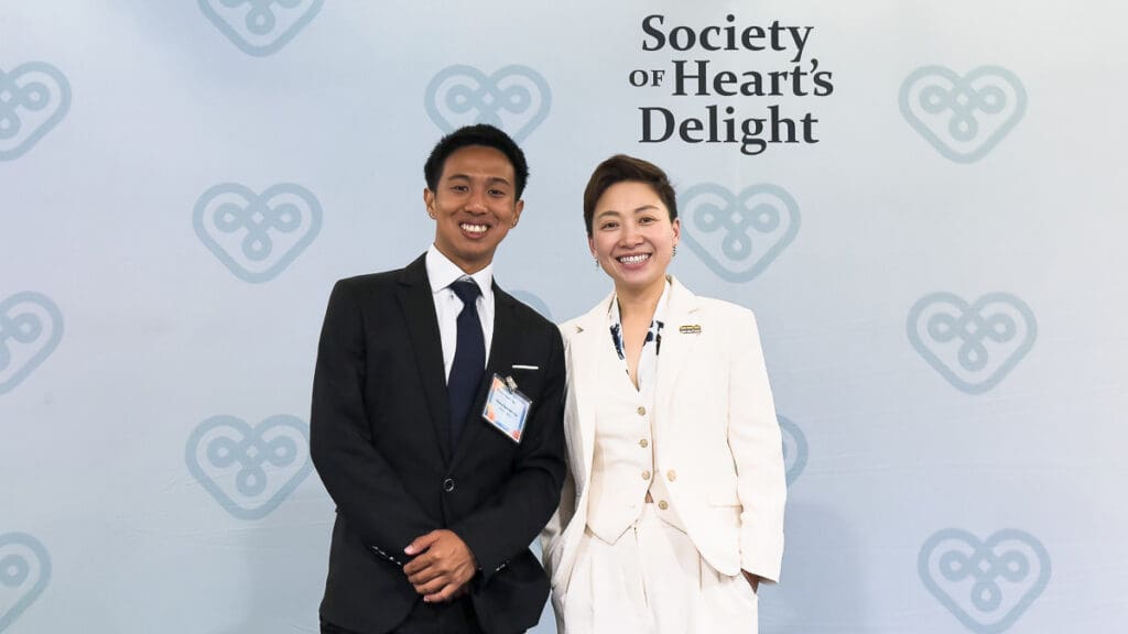 Two people stand smiling against a backdrop with heart motifs and "Society of Heart's Delight" text. They wear formal suits, radiating happiness.