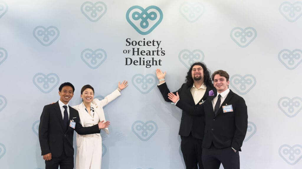 Four people stand smiling against a backdrop with heart motifs and "Society of Heart's Delight" text. They wear formal suits, radiating happiness.