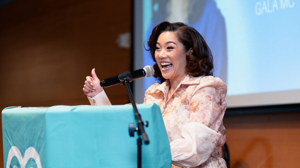A woman in a floral outfit stands behind a podium with a microphone, smiling widely and giving a thumbs up. The setting appears to be a formal event.