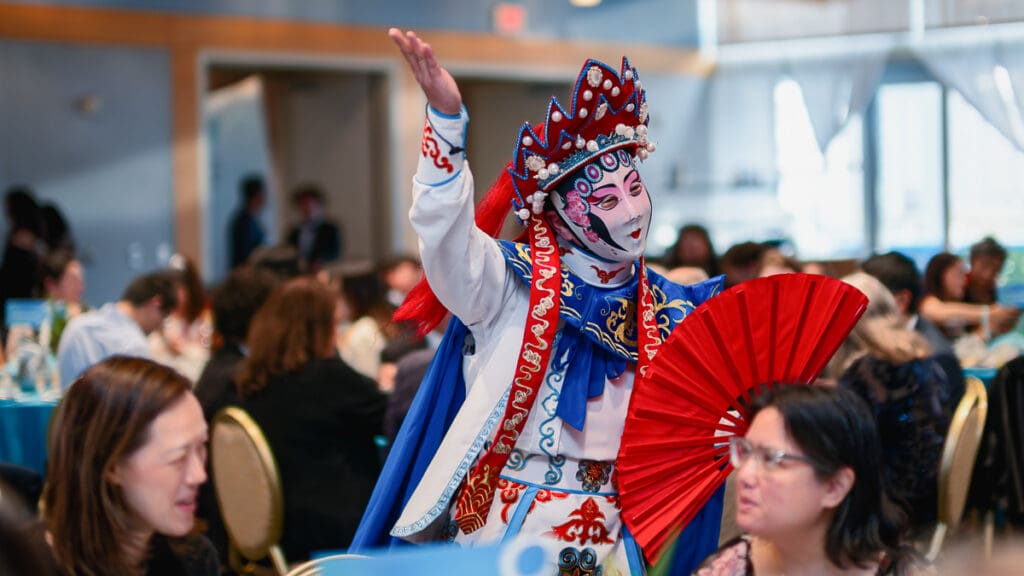 A performer in vibrant traditional Chinese opera attire, holding a red fan, engages with a seated audience. The atmosphere is lively and cultural.