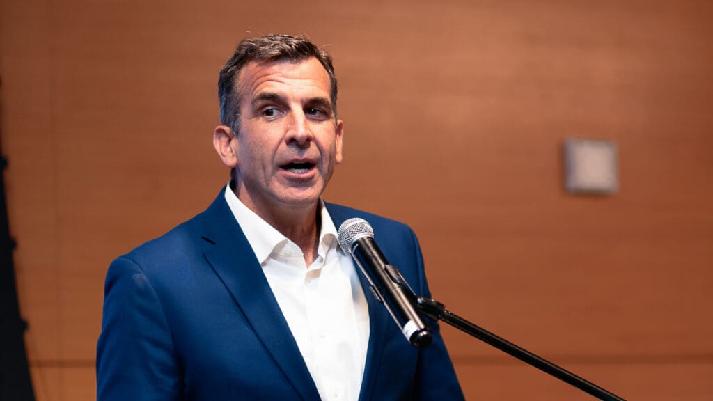 A man in a blue suit speaks into a microphone against a wood-paneled background, conveying confidence and engagement.