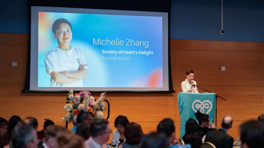 A speaker stands at a podium addressing an audience. A large screen shows “Michelle Zhang, Society of Heart's Delight, Founder & CEO.” The setting is a formal event.