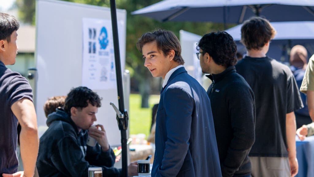 A young man in a blue suit stands outdoors at an event booth, surrounded by casually dressed people. He looks engaged, with a focused expression.