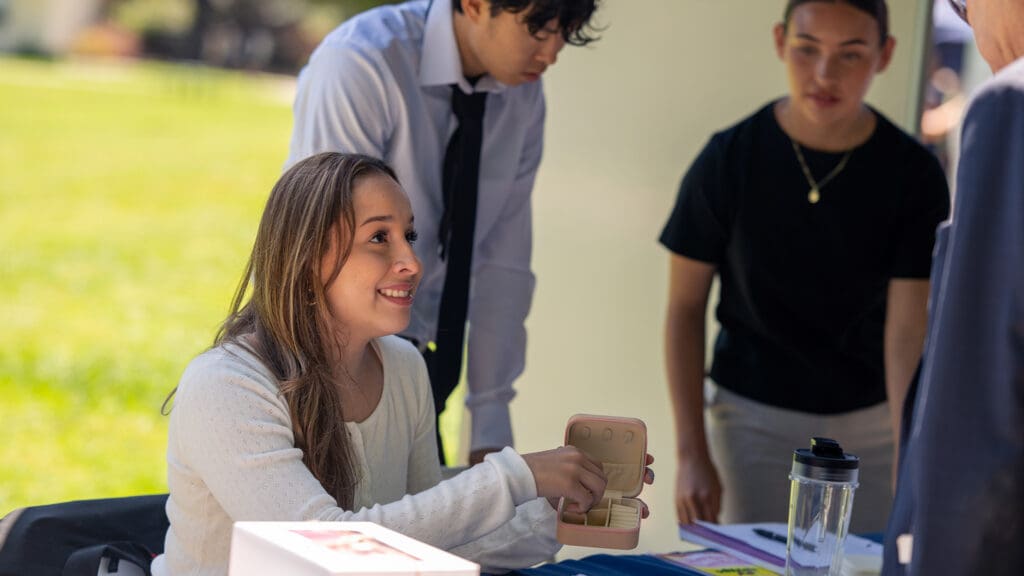 A woman at a table outdoors smiles while pointing to a small box. Two people stand nearby, looking on attentively. The scene is bright and engaging.