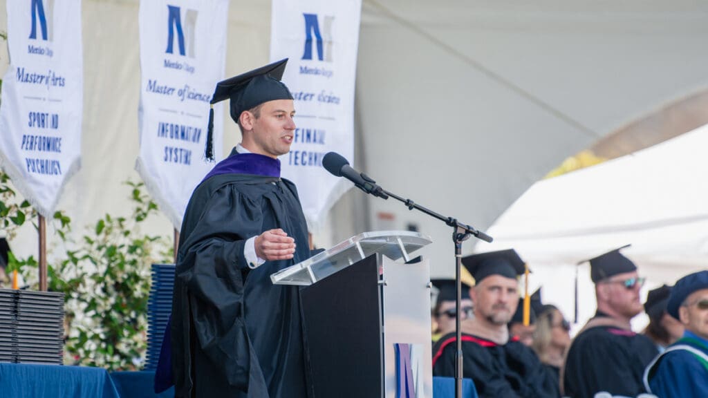 A man in graduation attire delivers a keynote address while other adults on the stage listen.