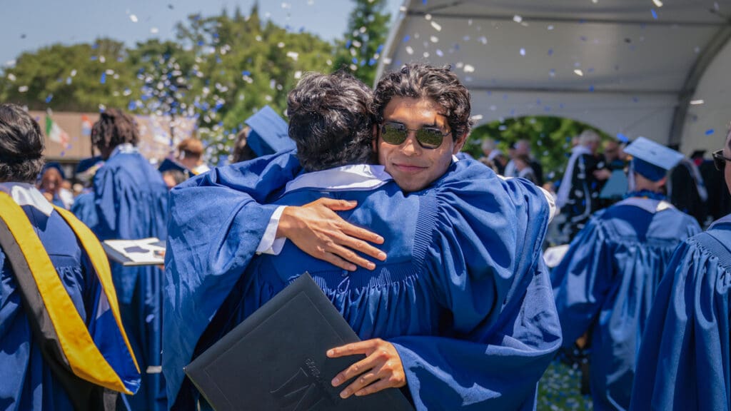 Graduates in blue gowns embrace joyfully at an outdoor ceremony. One holds a diploma with confetti falling around them, conveying celebration and happiness.