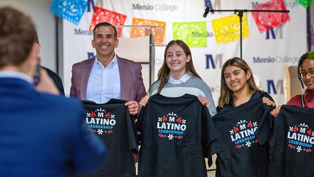 Three people stand together holding Latino Leadership Program T-shirts while posing for a photo in front of a Menlo College backdrop decorated with colorful banners.