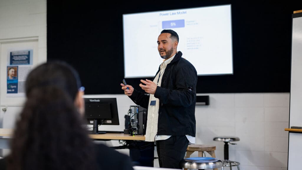 A man stands in a classroom setting, presenting to an audience. He is gesturing with a pen towards a projected slide on the wall titled "Power Law Model." The atmosphere is professional and focused.