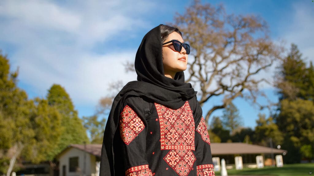 A person stands outdoors on the Menlo College campus wearing a black headscarf and a traditional embroidered dress, looking into the distance with trees and campus buildings in the background.