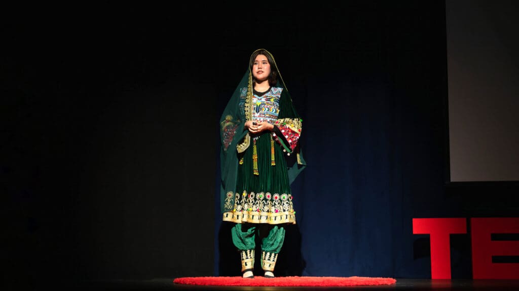 A person wearing a traditional green embroidered outfit stands on a stage speaking during a TEDx presentation, with a dark backdrop and red TEDx letters visible beside them.