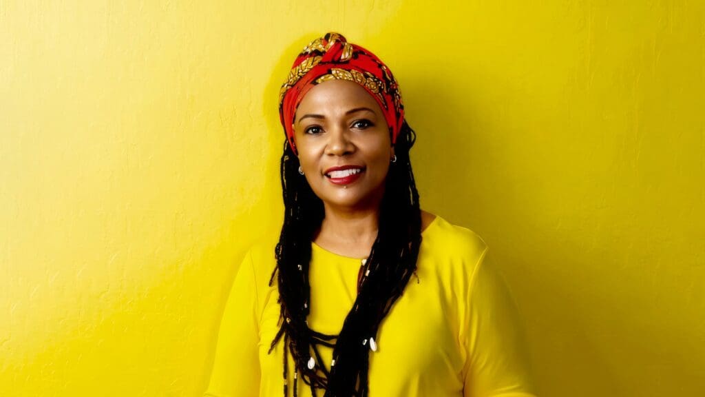Portrait of Menlo College lecturer Lakiba Pittman smiling against a bright yellow background, wearing a yellow blouse and a red patterned headwrap.