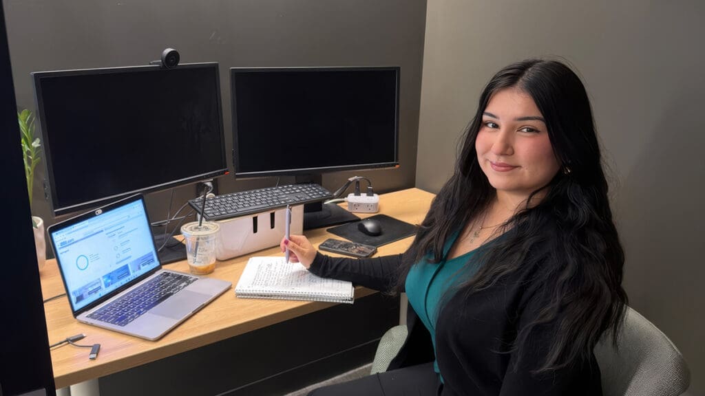 Menlo College student Jessica Oseguera Santoyo sits at a desk with a laptop, monitors, and notebook during her summer internship at Northwestern Mutual.
