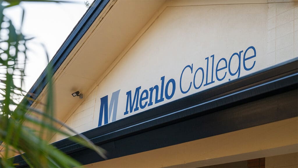 Exterior view of a Menlo College campus building featuring the blue Menlo College name and logo prominently displayed above the entrance, framed by greenery in the foreground.